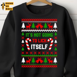 Op de afbeelding: Zwarte sweatshirt met een feestelijk kerstontwerp. Het ontwerp bevat rode rendieren, groene kerstbomen en de tekst "IT'S NOT GOING TO LICK ITSELF" in wit en groen. Zuurstokken zijn ook aanwezig.
