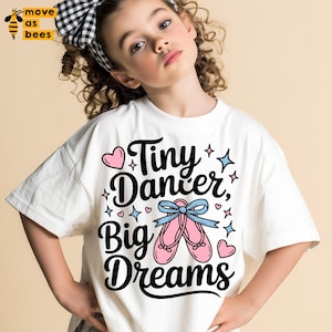 May include: White t-shirt with the text "Tiny Dancer, Big Dreams" in black script, featuring pink ballet shoes with a blue bow. The design includes pink hearts and blue stars. The shirt is worn by a young person.