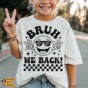 May include: A light beige t-shirt featuring a black graphic of a smiling emoji wearing sunglasses and making peace signs. The text "BRUH WE BACK!" is above and below the emoji, with a checkered pattern at the bottom.