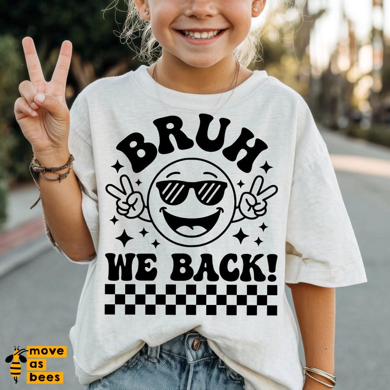 May include: A light beige t-shirt featuring a black graphic of a smiling emoji wearing sunglasses and making peace signs. The text "BRUH WE BACK!" is above and below the emoji, with a checkered pattern at the bottom.