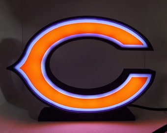 May include: Illuminated orange and purple "C" logo sign with a black border. The sign is lit with a bright orange centre and a purple outline. The sign is mounted on a black base and is likely a team logo.