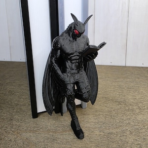 May include: A black 3D printed figurine of a muscular humanoid moth creature with large wings, pointed ears, and bright red eyes. The figure is depicted in a standing pose, holding and reading a small open book, with detailed textures on the body.