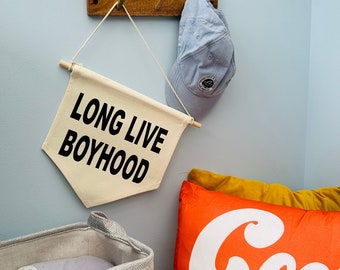 May include: A white canvas banner with the text "Long Live Boyhood" hanging on a wooden hook. A light blue baseball cap hangs on the hook next to the banner. A gray storage bin with a rocket ship design sits on the floor. A red pillow with the text "Cool to be Kind" in white letters sits on a yellow surface.