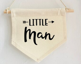 May include: A cream-colored banner with the words "LITTLE Man" in black script and an arrow. The banner is suspended by a wooden dowel and a natural-colored cord, suitable for nursery decor.