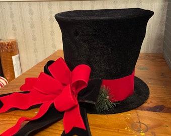 May include: A black velvet top hat with a red velvet ribbon and a red bow. The hat is decorated with a small sprig of pine.