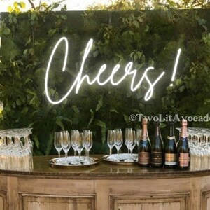 Peut inclure: Un bar en bois avec un mur vert derri&egrave;re. Le mur a une enseigne au n&eacute;on qui dit "Cheers!" et le texte "&copy;TwoLitAvocados". Le bar est d&eacute;cor&eacute; de bouteilles de champagne et de verres.