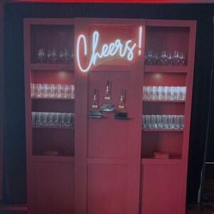 Peut inclure: Un bar en bois rouge avec des &eacute;tag&egrave;res remplies de verres et un n&eacute;on qui dit "Cheers!" Un tapis &agrave; motif de peau de tigre se trouve devant le bar.