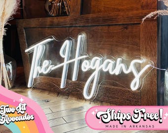 May include: A custom neon sign featuring the white illuminated script text The Hogans mounted on a clear acrylic backing with a wavy edge. The sign is positioned against a dark wood surface, displaying bright white light in a cursive font style.