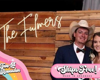 May include: A custom neon sign featuring the text The Fulmers in a cursive script, mounted on a horizontal wooden plank wall. The sign emits a bright white light, creating a focal point against the natural brown wood texture of the backdrop.