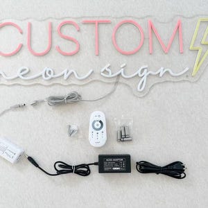 Peut inclure: Un panneau lumineux n&eacute;on blanc avec les mots "CUSTOM neon sign" et un &eacute;clair jaune. Le panneau est sur une surface blanche avec une t&eacute;l&eacute;commande blanche, un adaptateur secteur et un cordon d'alimentation.