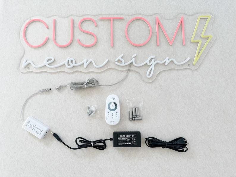 Peut inclure: Un panneau lumineux n&eacute;on blanc avec les mots "CUSTOM neon sign" et un &eacute;clair jaune. Le panneau est sur une surface blanche avec une t&eacute;l&eacute;commande blanche, un adaptateur secteur et un cordon d'alimentation.