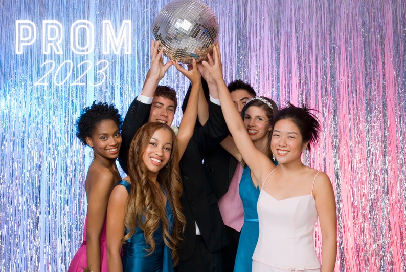 Puede incluir: Un grupo de seis j&oacute;venes posan para una foto frente a un tel&oacute;n de fondo brillante plateado y rosa. Est&aacute;n sosteniendo una gran bola de discoteca sobre sus cabezas. El texto "PROM 2023" es visible en el fondo.