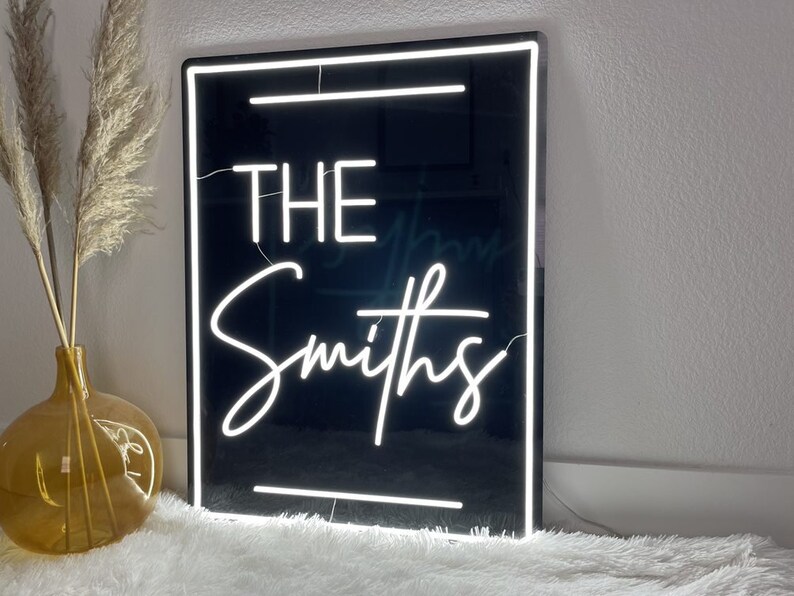 Puede incluir: Un letrero de ne&oacute;n negro con letras de ne&oacute;n blancas que dicen "THE Smiths". El letrero est&aacute; enmarcado con un borde blanco y tiene un fondo negro.