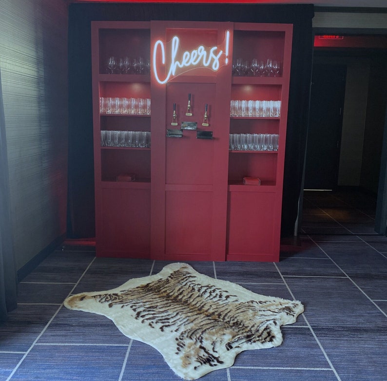 Peut inclure: Un bar en bois rouge avec des &eacute;tag&egrave;res remplies de verres et un n&eacute;on qui dit "Cheers!" Un tapis &agrave; motif de peau de tigre se trouve devant le bar.