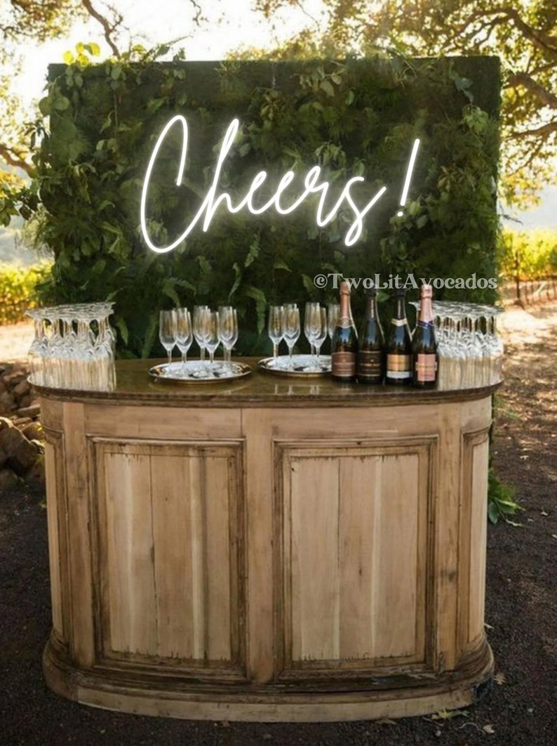 Peut inclure: Un bar en bois avec un mur vert derri&egrave;re. Le mur a une enseigne au n&eacute;on qui dit "Cheers!" et le texte "&copy;TwoLitAvocados". Le bar est d&eacute;cor&eacute; de bouteilles de champagne et de verres.