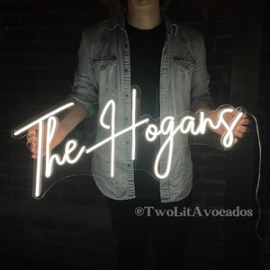 Peut inclure: Un panneau lumineux blanc en n&eacute;on avec le texte "The Hogans" en &eacute;criture cursive. Le panneau est tenu par une personne portant une veste en jean.
