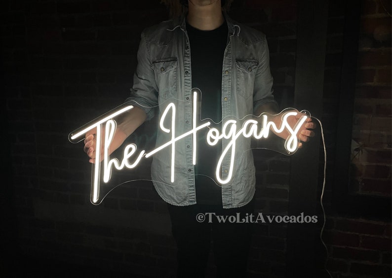 Peut inclure: Un panneau lumineux blanc en n&eacute;on avec le texte "The Hogans" en &eacute;criture cursive. Le panneau est tenu par une personne portant une veste en jean.