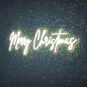 Peut inclure: Un panneau lumineux blanc en n&eacute;on avec les mots "Merry Christmas" sur un fond de verdure.