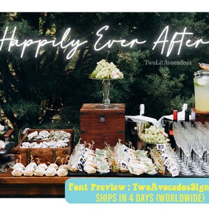 May include: A white neon sign with the text "Happily Ever After" hanging above a table with food and drinks. The table is set up for a wedding reception or party. There are also two wooden chests on the table.