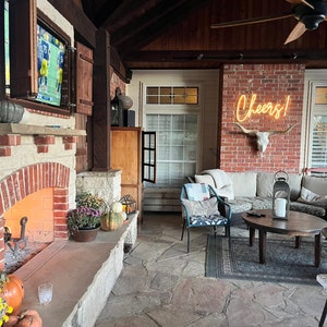 Peut inclure: Une terrasse en briques avec une chemin&eacute;e en pierre, un ventilateur de plafond et une enseigne au n&eacute;on qui dit "Cheers!" Il y a un canap&eacute;, une table basse et une chaise avec un coussin qui dit "Be Our Guest".