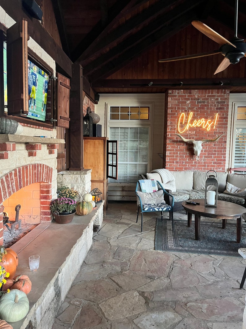 Peut inclure: Une terrasse en briques avec une chemin&eacute;e en pierre, un ventilateur de plafond et une enseigne au n&eacute;on qui dit "Cheers!" Il y a un canap&eacute;, une table basse et une chaise avec un coussin qui dit "Be Our Guest".