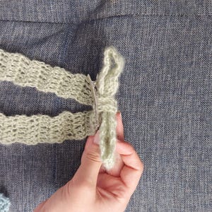 May include: A light green crocheted bow with a silver clip. The bow is held in a hand.