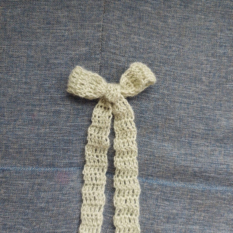 May include: A beige crocheted bow with two long tails on a blue textured background.