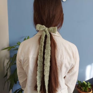 May include: A green crocheted hair tie in the shape of a bow. The hair tie is tied around a ponytail.