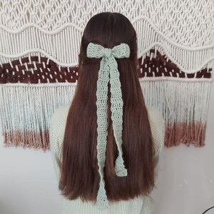May include: A light green crocheted hair bow with long tails. The bow is tied in a knot at the top and the tails are hanging down.