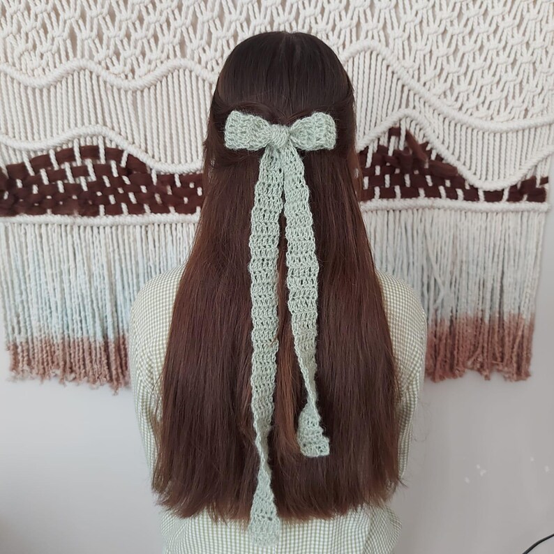 May include: A light green crocheted hair bow with long tails. The bow is tied in a knot at the top and the tails are hanging down.
