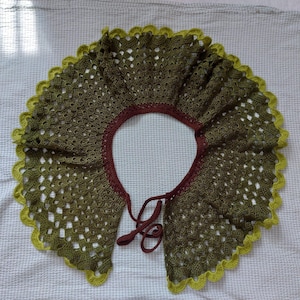 Puede incluir: Un collar de ganchillo en tonos verdes y marrones con un borde festoneado. El collar tiene un cierre de corbata.