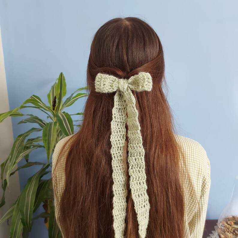 May include: A light green crocheted bow hair clip with long tails. The bow is tied in a simple knot and the tails are long and flowing.