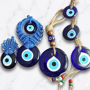 May include: Two blue macrame wall hangings featuring multiple dark blue glass evil eye beads. The left piece has intricate blue cord knotting, while the right piece uses natural jute twine with wooden beads and smaller colorful accent beads.