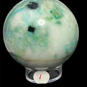 May include: A polished spherical stone featuring a marbled appearance with white, light blue, and green hues, accented by small dark black inclusions. The sphere is displayed on a clear, circular acrylic stand with a white sticker labeled with the number 1.