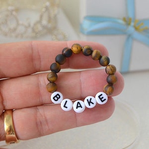 May include: A brown and gold tiger's eye beaded bracelet with white beads spelling out the name "BLAKE".