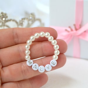 Peut inclure: Un bracelet de perles avec le nom "MAEVE" épelé en perles de lettres blanches. Le bracelet est tenu dans une main, avec une boîte cadeau blanche attachée avec un ruban rose et un diadème argenté en arrière-plan.