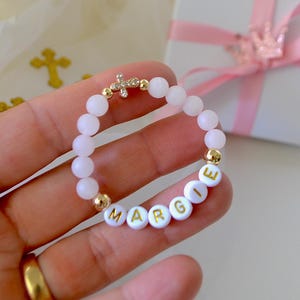 Puede incluir: Una pulsera de cuentas rosa claro con el nombre "MARGIE" escrito con cuentas de letras blancas. La pulsera presenta detalles dorados y un pequeño dije de cruz. En el fondo, una caja de regalo blanca con una cinta rosa.