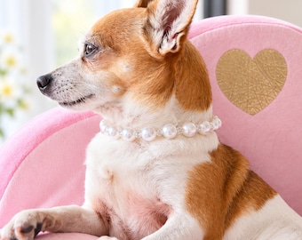 May include: A small brown and white dog wearing a white pearl collar sits on a pink upholstered pet sofa featuring a gold heart emblem. A white square gift box tied with a pink satin ribbon rests beside the dog on the pink cushion.