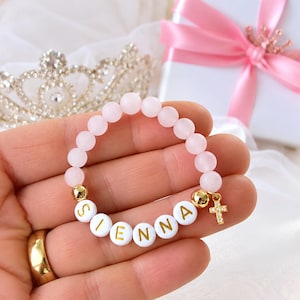 May include: A stretch bracelet featuring matte light pink beads, white circular beads with gold lettering spelling the name SIENNA, gold-toned spacer beads, and a small gold-toned cross charm with embedded clear stones.