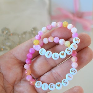May include: Two beaded bracelets with white beads and gold lettering. The larger bracelet says "BIG SIS" and the smaller bracelet says "LIL SIS". Both bracelets have pink and yellow beads.