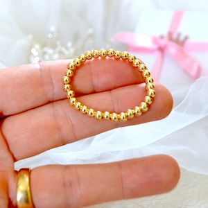 May include: A gold beaded ring, composed of small, round beads, is held in a hand. The ring is a bright, shiny gold color. A gold ring is visible on the finger of the hand. A white gift box with a pink ribbon is in the background.