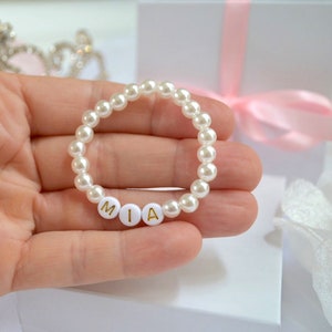Puede incluir: Un brazalete de perlas blancas con letras doradas que deletrean el nombre "MIA".