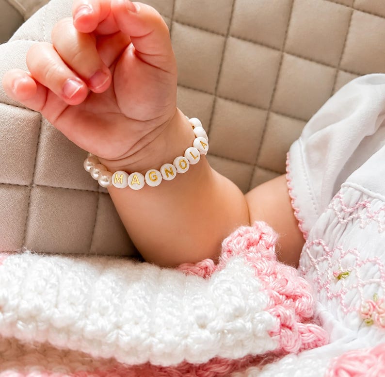 Puede incluir: Una pulsera de cuentas con esferas blancas y cuentas redondas blancas con letras doradas que forman la palabra MAGNOLIA. La pulsera est&aacute; compuesta por una alternancia de cuentas tipo perla y cuentas con letras en forma circular.