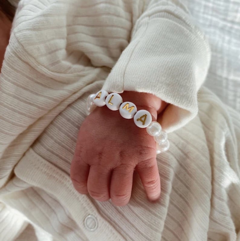 Puede incluir: Una pulsera con cuentas esf&eacute;ricas blancas y cuentas circulares blancas con las letras doradas A, L, M y A impresas. La pulsera se lleva en la mu&ntilde;eca, con las cuentas rodeando la mu&ntilde;eca sobre una tela de color crema.