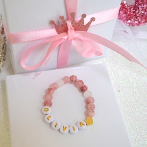 Pode incluir: Um bracelete de contas rosa e branco com o nome "Olivia" escrito em letras brancas com detalhes dourados. O bracelete tem um pequeno pingente de cora&ccedil;&atilde;o dourado.