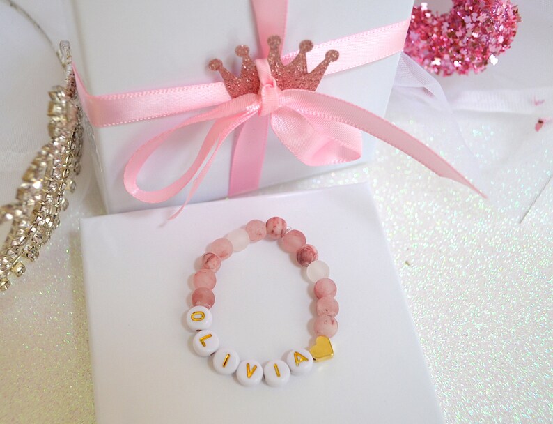 Pode incluir: Um bracelete de contas rosa e branco com o nome "Olivia" escrito em letras brancas com detalhes dourados. O bracelete tem um pequeno pingente de cora&ccedil;&atilde;o dourado.