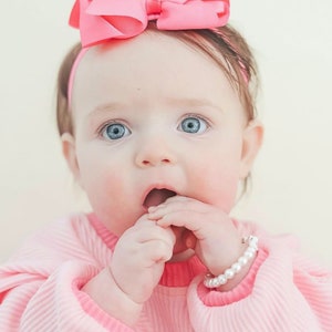 Puede incluir: Un lazo rosa para el pelo sobre una diadema fina, una camiseta de manga larga acanalada de color rosa claro con cuello de un rosa m&aacute;s oscuro y una pulsera de perlas blancas de imitaci&oacute;n en la mu&ntilde;eca. Los accesorios y la ropa se ven claramente.