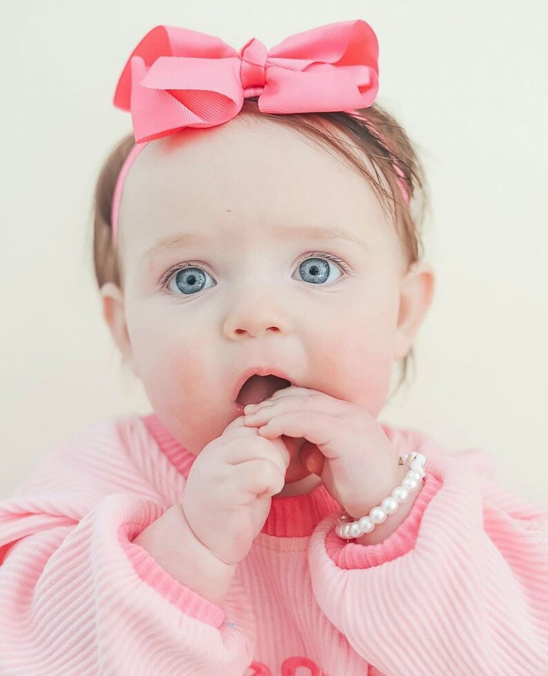 Puede incluir: Un lazo rosa para el pelo sobre una diadema fina, una camiseta de manga larga acanalada de color rosa claro con cuello de un rosa m&aacute;s oscuro y una pulsera de perlas blancas de imitaci&oacute;n en la mu&ntilde;eca. Los accesorios y la ropa se ven claramente.