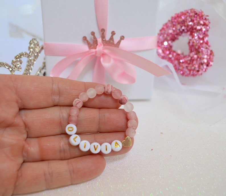 Pode incluir: Um bracelete de contas rosa e branco com o nome "Olivia" escrito em letras douradas. O bracelete tem um pequeno charme de cora&ccedil;&atilde;o dourado.