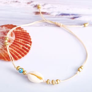 May include: Anklet featuring a natural seashell, a blue evil eye bead, and gold-colored beads strung on a beige cord. The adjustable cord allows for a comfortable fit. This beach-themed jewelry is perfect for summer.